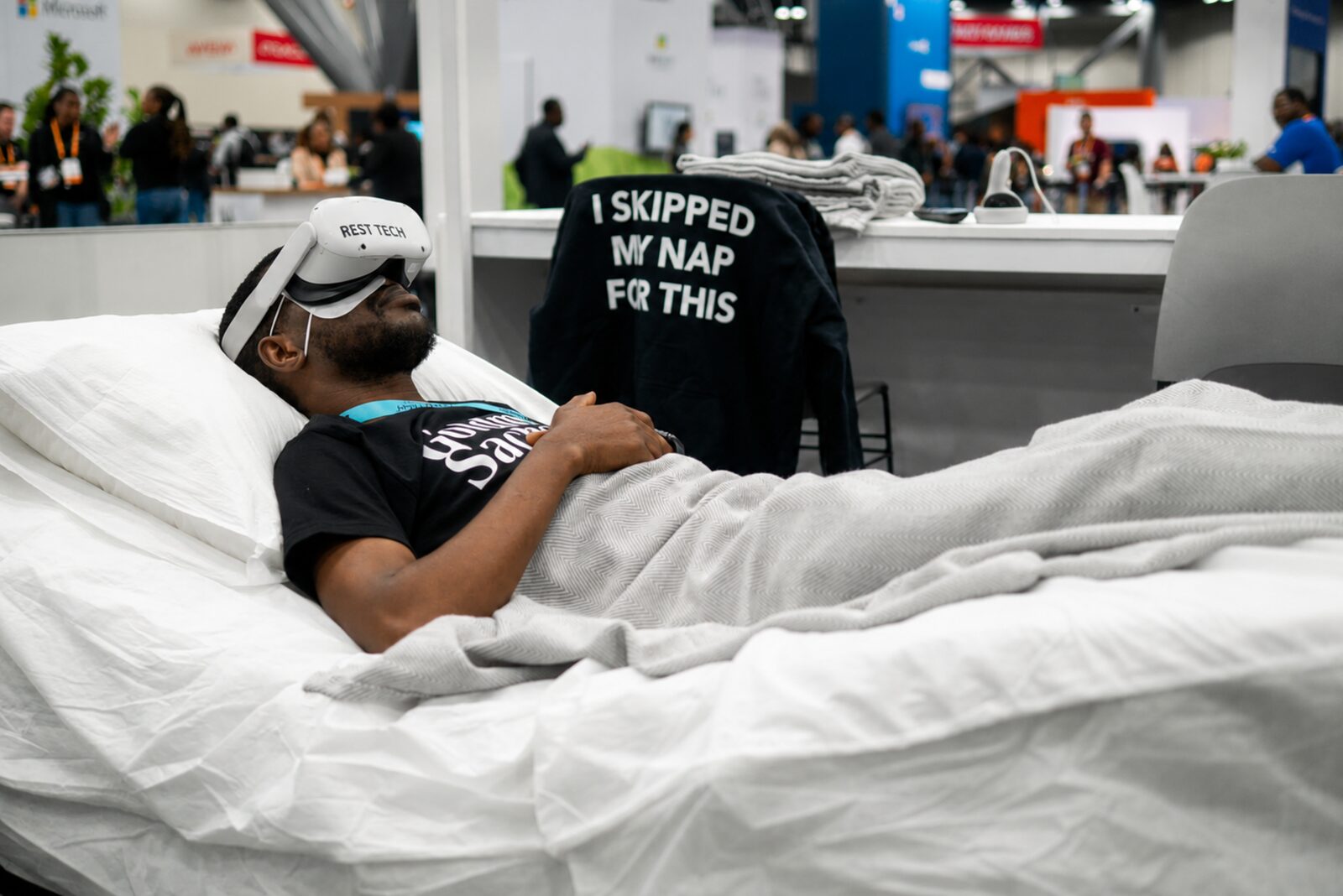 A conference attendee resting on a bed wearing a white Rest Tech VR headset, an “I skipped my nap for this” jacket draped on the chair beside him.